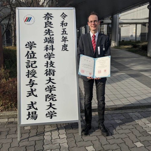 Johannes hält sein Abschlusszeugnis in der Hand und steht neben einem großen Schild mit japanischen Zeichen.